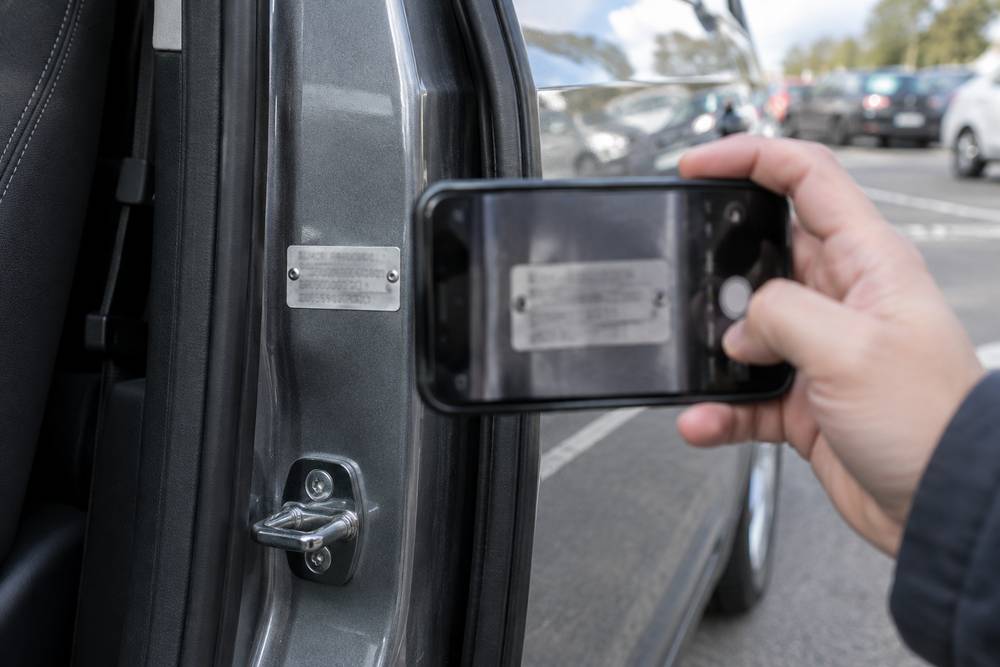 Main photographiant au smartphone la plaque constructeur rivetée sur le montant de porte d'une voiture grise