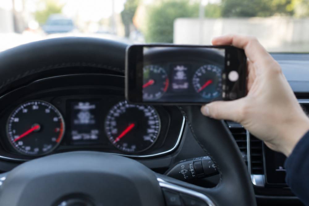 Main tenant un smartphone devant le combiné d'instruments d'une voiture pour photographier le compteur, volant au premier plan
